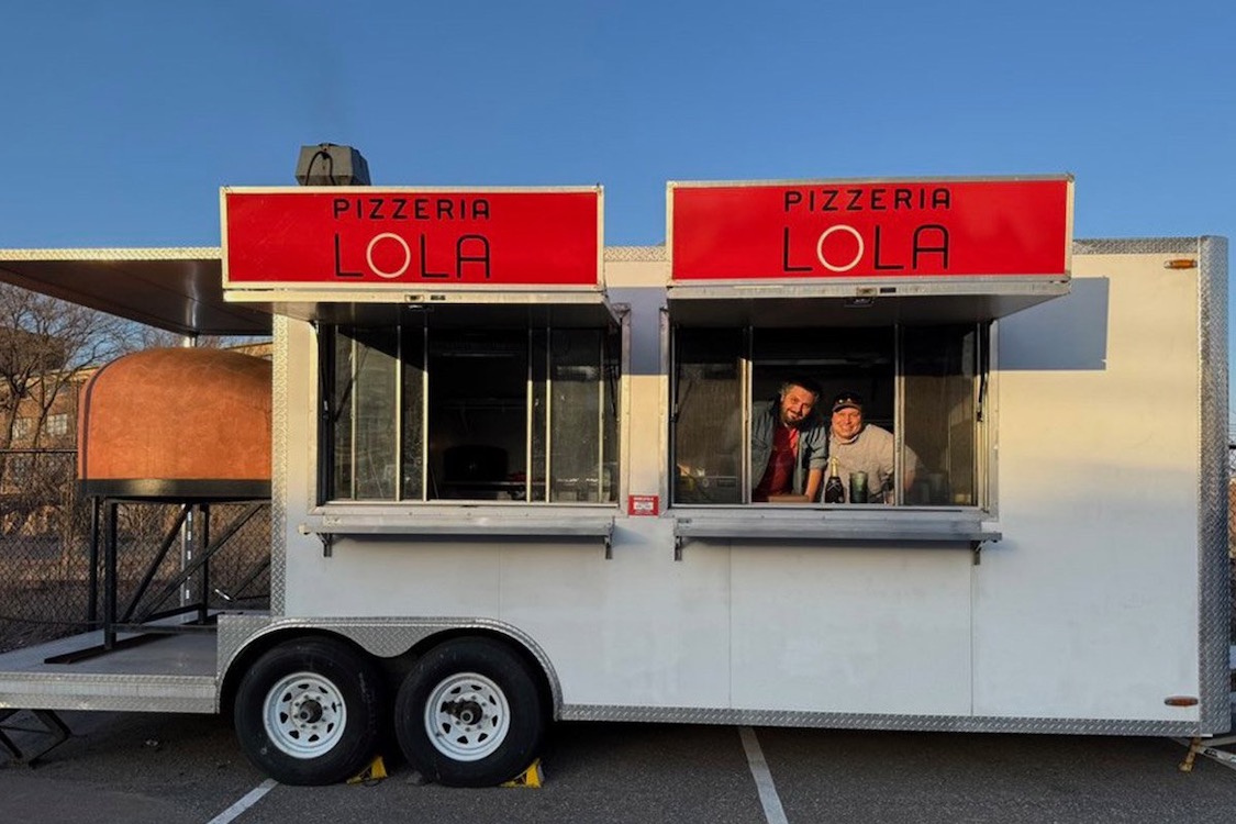 This photo shows two men poking their heads out of one of the windows in the Pizzeria Lola food trailer, with two Pizzeria Lola signs on top of the trailer and a dome-shaped wood fired oven visible on the far left.