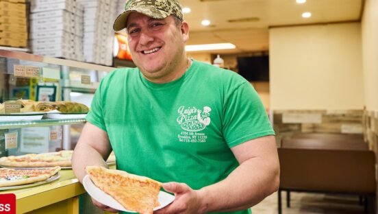 This photo shows a man in a green Luigi's Pizza t-shirt holding a cheese slice in a paper plate.