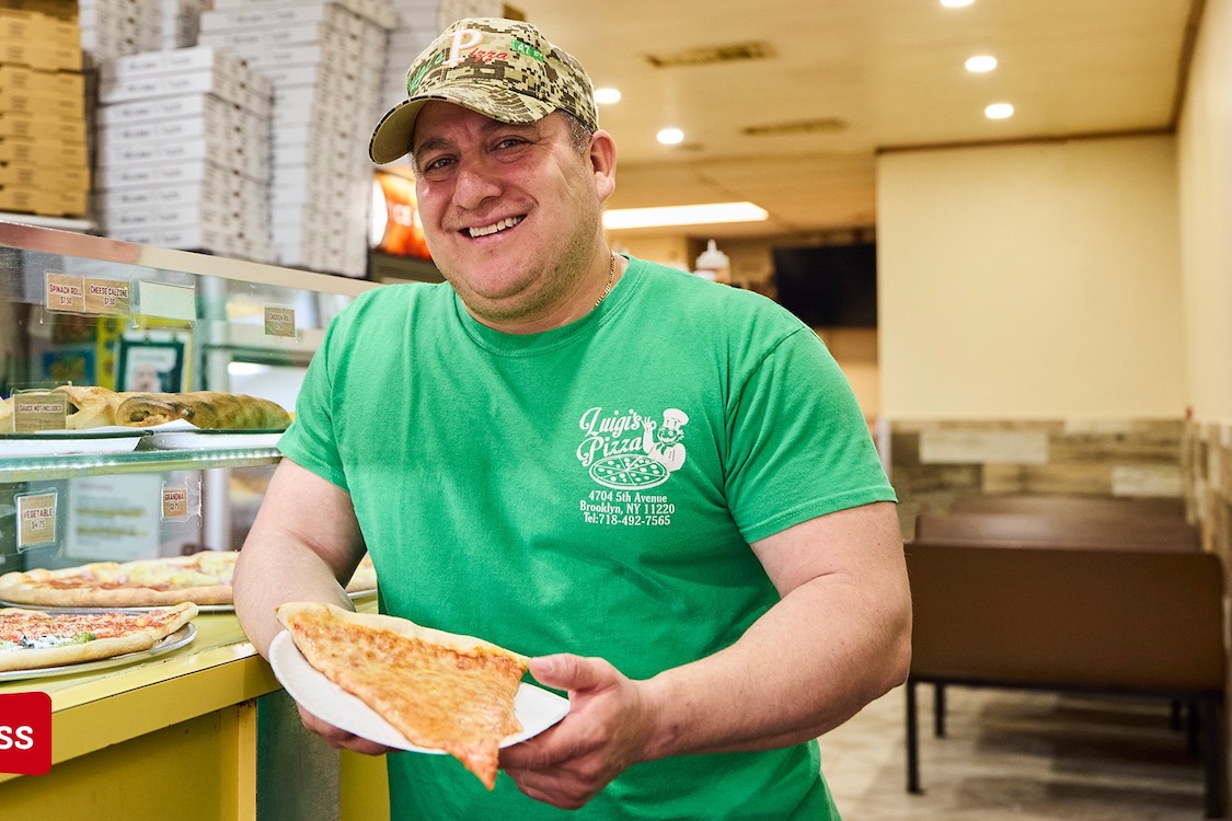 This photo shows a man in a green Luigi's Pizza t-shirt holding a cheese slice in a paper plate.