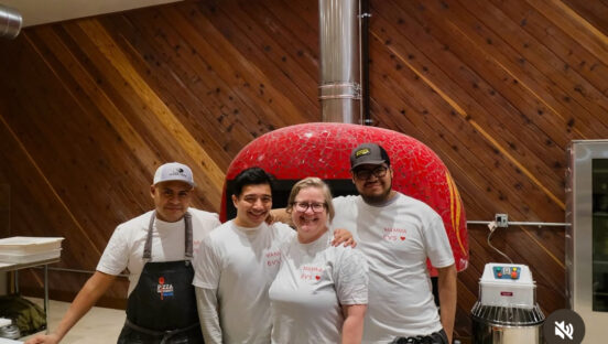 This photo shows Mama Ev's owner and three men, all wearing branded t-shirts, standing in front of a wood-fired oven with a large red dome..