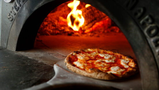 This photo shows a Neapolitan pizza topped with mozzarella on a peel in front of a wood-burning oven.