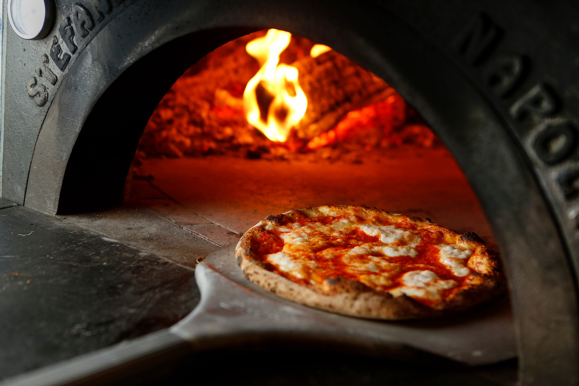This photo shows a Neapolitan pizza topped with mozzarella on a peel in front of a wood-burning oven.