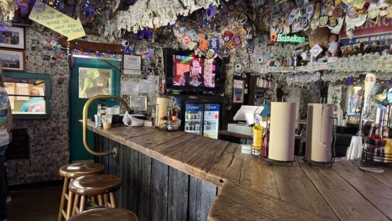 This photo shows the rustic interior of No Name Pub with a bar made of wood and an overhead string with dozens of what look to be dollar bills and more bills lining the walls.