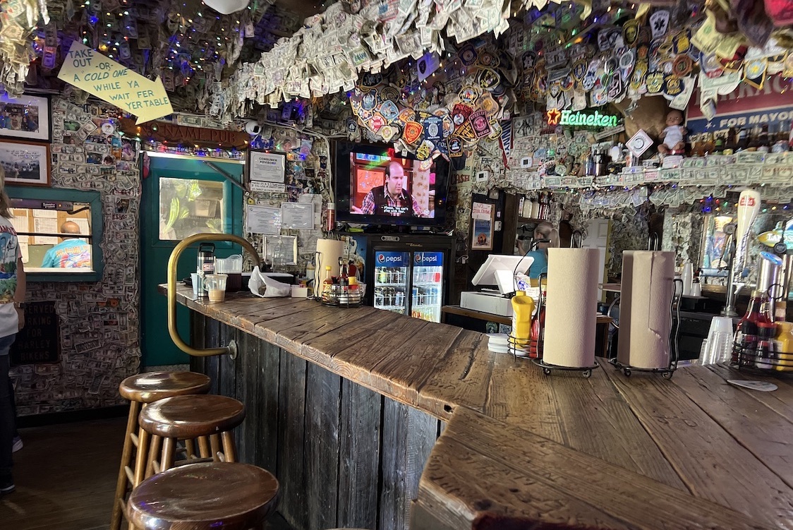 This photo shows the rustic interior of No Name Pub with a bar made of wood and an overhead string with dozens of what look to be dollar bills and more bills lining the walls.