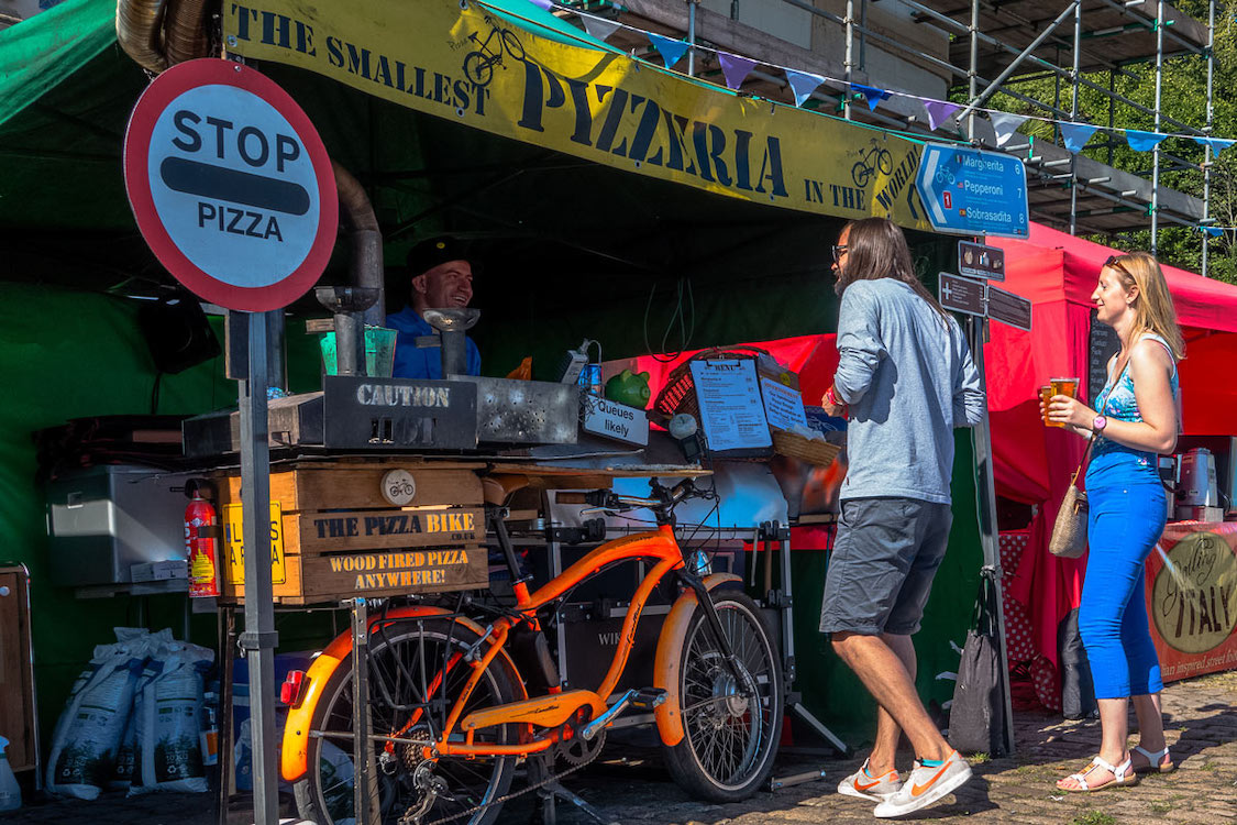 This photo shows The Pizza Bike's tent at a festival with the bike parked in front and a yellow sign with black type that reads, "The Smallest Pizzeria in the World."