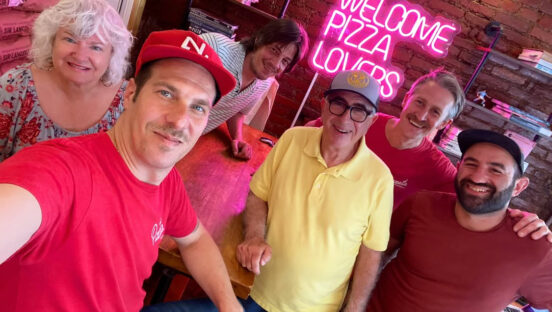 This photo shows Mary Ann and Paulie Gee posing for a photo with the staff at Pizzata Pizzeria in Philadelphia. Behind them is a red-neon sign that reads, "Welcome Pizza Lovers."