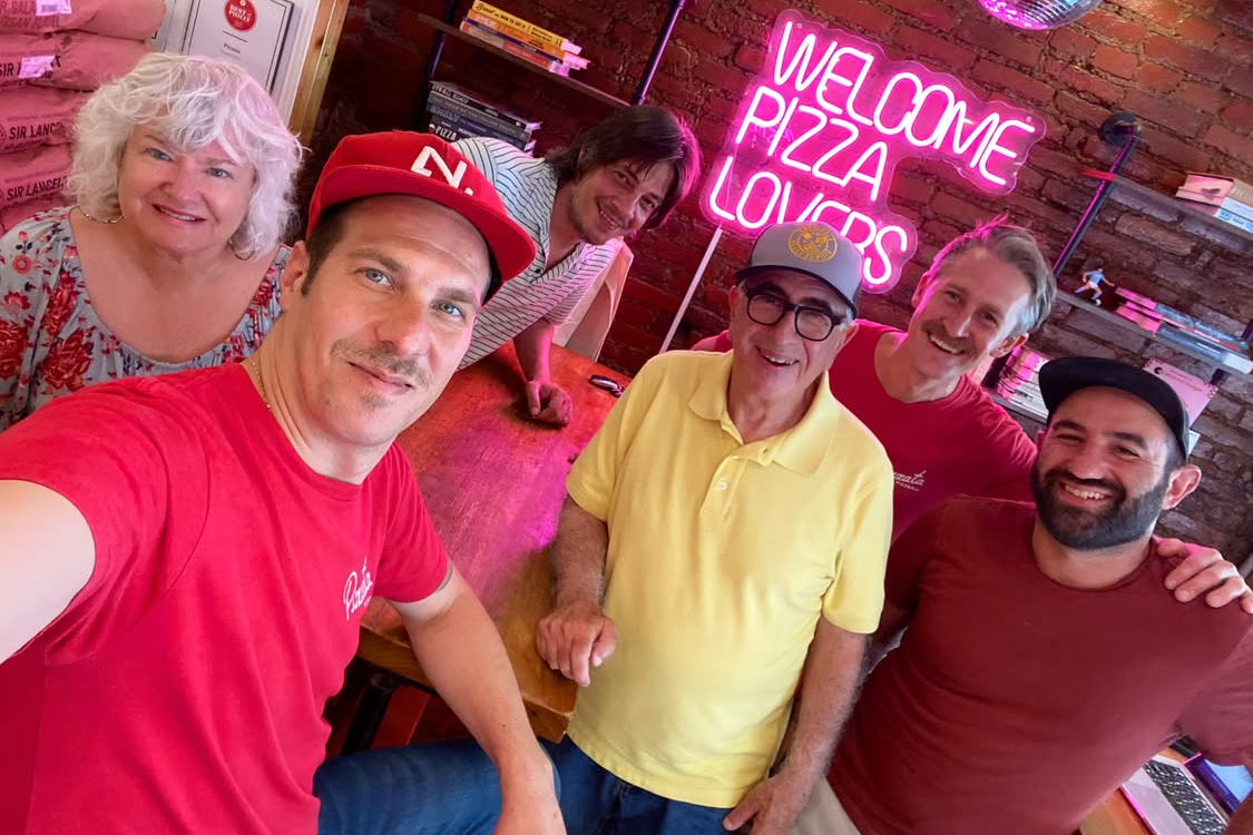This photo shows Mary Ann and Paulie Gee posing for a photo with the staff at Pizzata Pizzeria in Philadelphia. Behind them is a red-neon sign that reads, "Welcome Pizza Lovers."