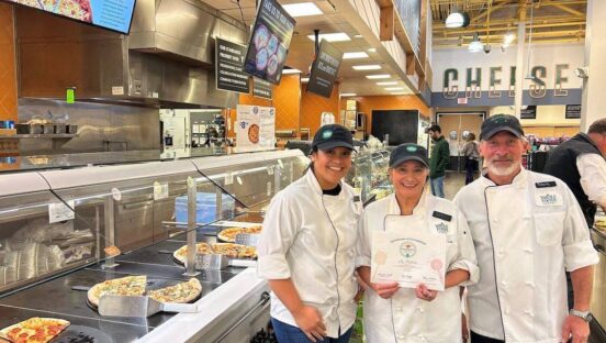 This photo shows three people - two women and one man - in white chef coats, standing in a Whole Foods store. The woman in the middle is holding a certificate.