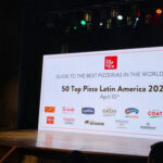 This photo shows a man standing at a podium on stage at 50 Top Pizza's awards presentation. Behind him is a huge screen with the words, "50 Top Pizza Latin America 2025"