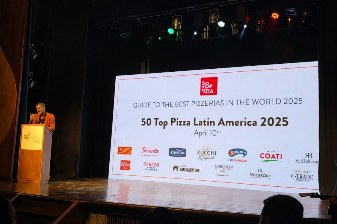 This photo shows a man standing at a podium on stage at 50 Top Pizza's awards presentation. Behind him is a huge screen with the words, "50 Top Pizza Latin America 2025"