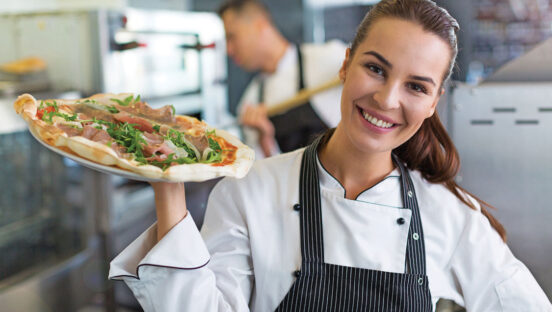 This photo shows a young woman in a white shirt and black apron, smiling and holding up a pizza near her shoulders.
