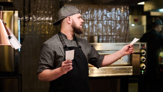 This photo shows a restaurant cook staring at printed orders or maybe receipts in the kitchen, as if studying them intently.