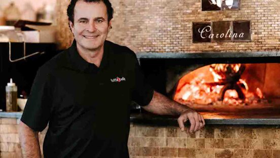 This photo shows Alex Camara in a black shirt, standing in front of a wood-fired oven.