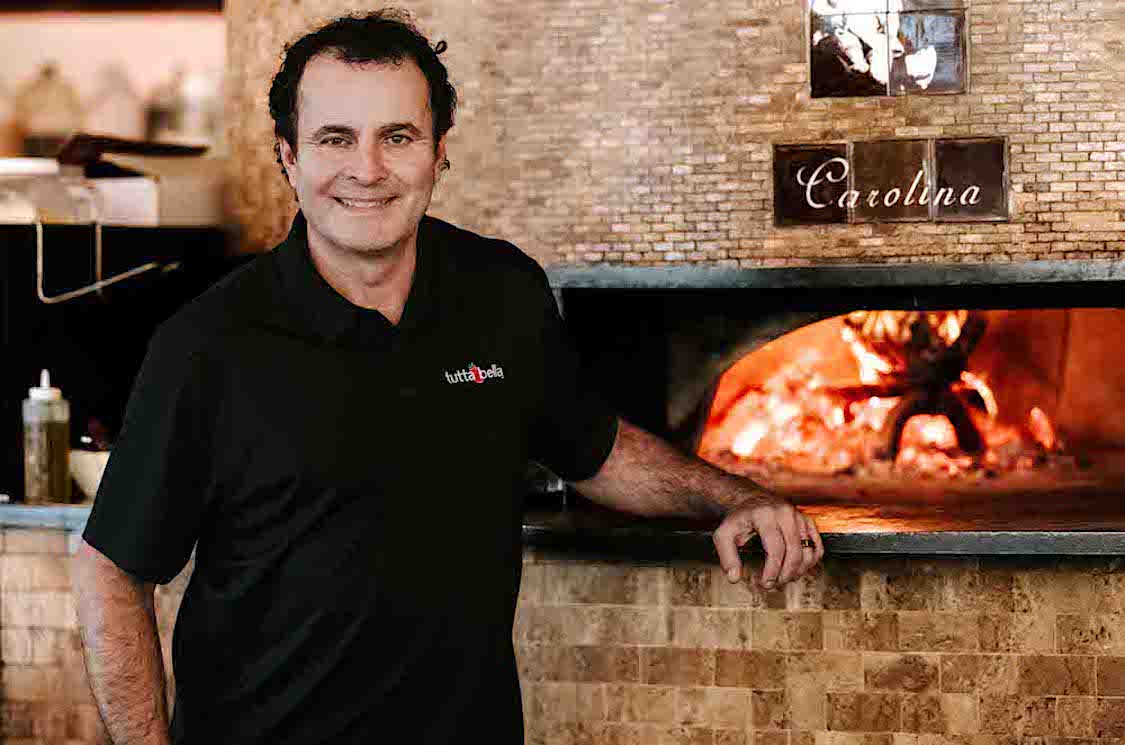 This photo shows Alex Camara in a black shirt, standing in front of a wood-fired oven.