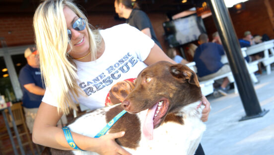 This photo shows a young woman with blonde hair and sunglasses, wearing a Cane Rosso Rescue t-shirt and hugging a brown and white dog.