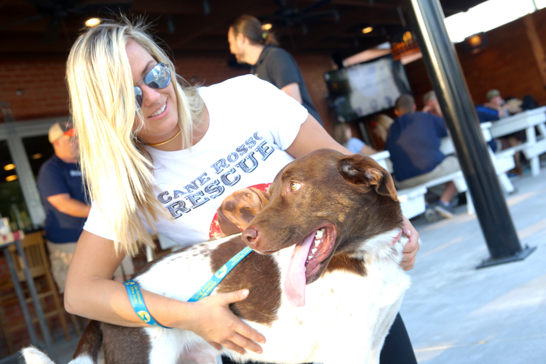 This photo shows a young woman with blonde hair and sunglasses, wearing a Cane Rosso Rescue t-shirt and hugging a brown and white dog.