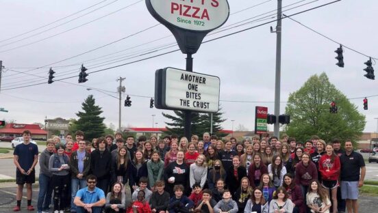This photo shows a group of high school students posing in front of the Cappy's Pizza sign outside. The sign reads, "Another One Bites the Crust."