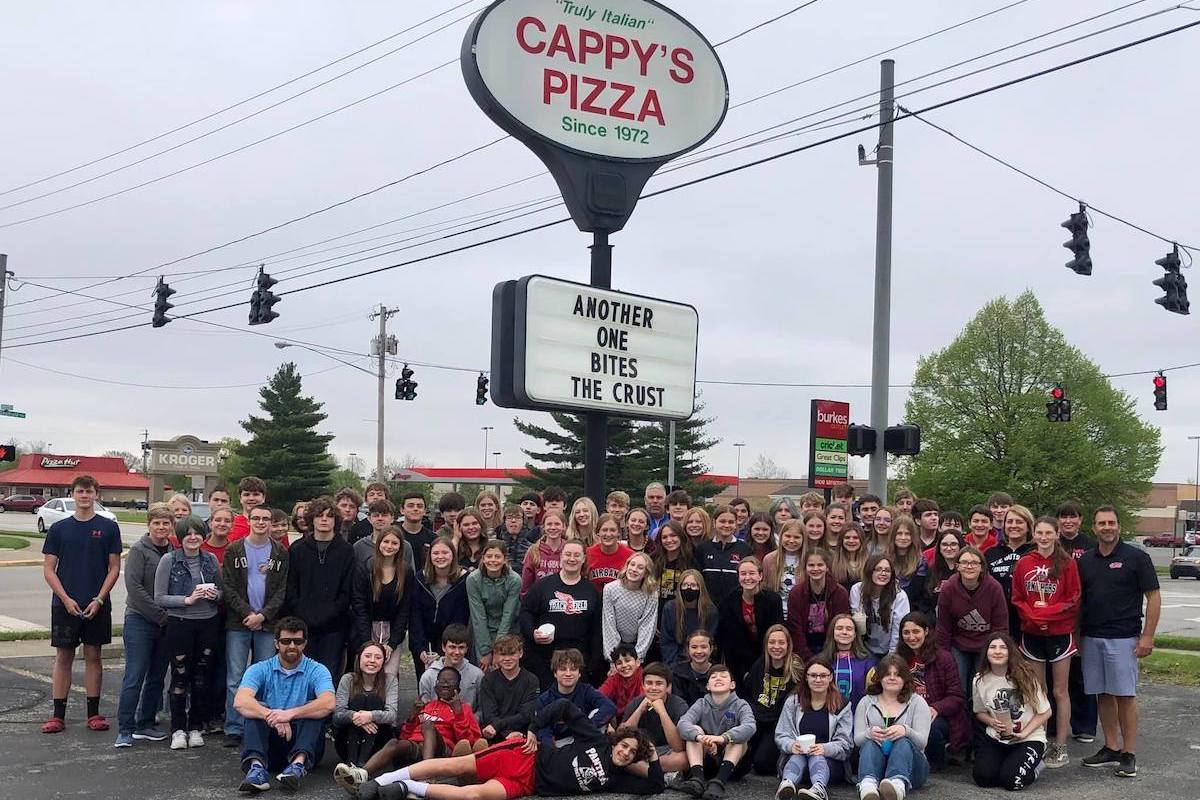 This photo shows a group of high school students posing in front of the Cappy's Pizza sign outside. The sign reads, "Another One Bites the Crust."