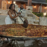 This photo shows a newlywed couple, both dressed in white, slicing into a gigantic 8-foot pizza with a huge slicer at their wedding.