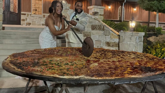 This photo shows a newlywed couple, both dressed in white, slicing into a gigantic 8-foot pizza with a huge slicer at their wedding.