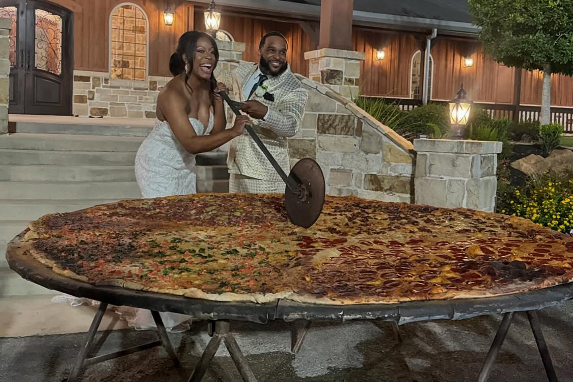 This photo shows a newlywed couple, both dressed in white, slicing into a gigantic 8-foot pizza with a huge slicer at their wedding.