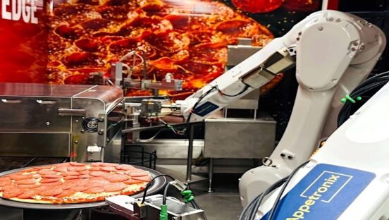 This photo shows a two-armed robot working with a pepperoni pizza, with a pizza oven in the background.