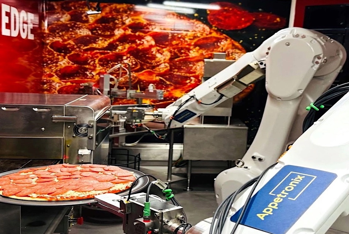 This photo shows a two-armed robot working with a pepperoni pizza, with a pizza oven in the background.
