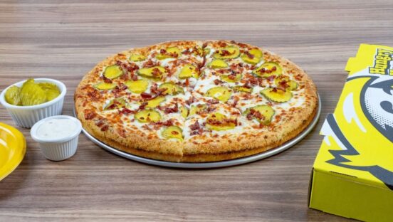 This photo shows Hungry Howie's Pickle Bacon Ranch Pizza next to a selection of dipping sauces and a carryout box.