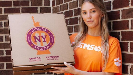 This photo shows a female employee in an orange Clemson shirt holding up a Joe's NY Pizza box.