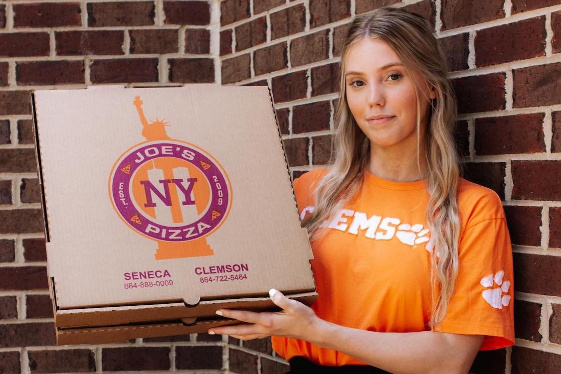 This photo shows a female employee in an orange Clemson shirt holding up a Joe's NY Pizza box.
