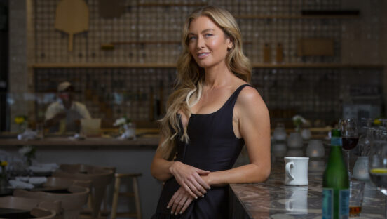 This photo shows Katie Lee in a glamorous black dress, leaning against the bar in one of her restaurants.