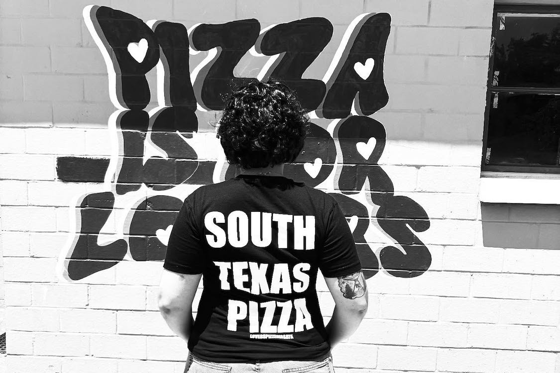 This black-and-white photo shows Lovers Pizzeria owner Victoria Moreno, back to camera, with a branded t-shirt that reads "South Texas Pizza." She is standing in front of a wall with lettering that reads, "Pizza Is For Lovers."