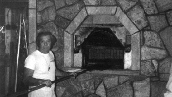 This vintage black-and-white photo shows a man in a white t-shirt standing in front of the pizza oven at Modern Apizza.