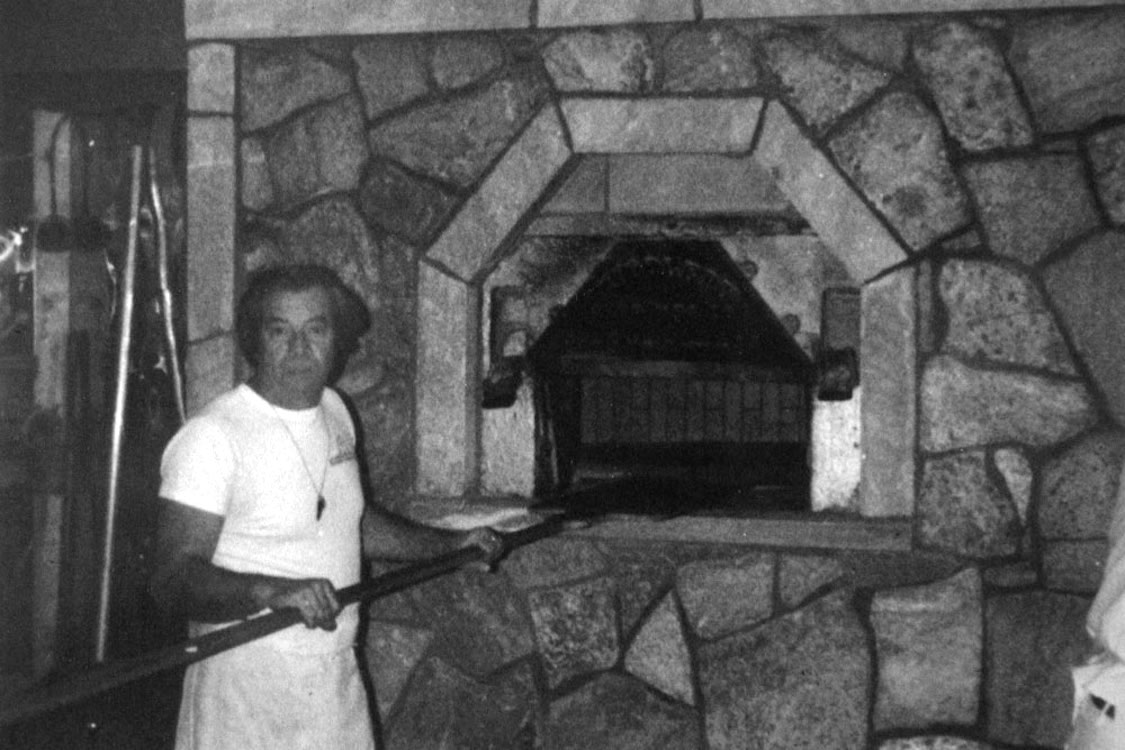 This vintage black-and-white photo shows a man in a white t-shirt standing in front of the pizza oven at Modern Apizza.
