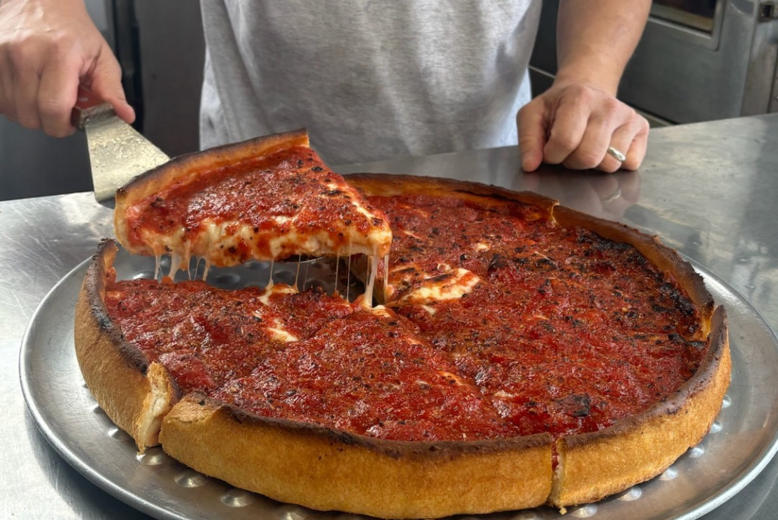 This photo shows a man's hand pulling out a slice of deep-dish pizza with a spatula.