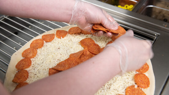 This photo shows a woman's hands, wearing gloves, adding pepperoni slices to an unbaked pizza.