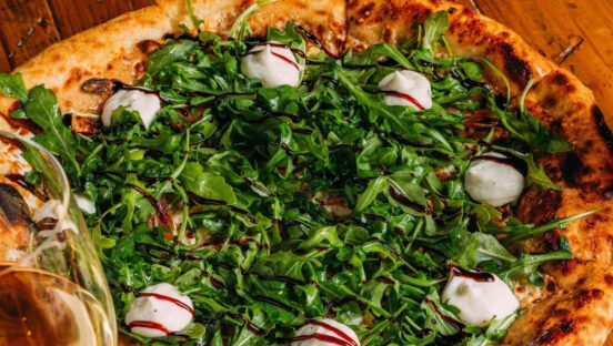 This photo shows a pizza topped with arugula, ricotta, prosciutto, dates and other ingredients.