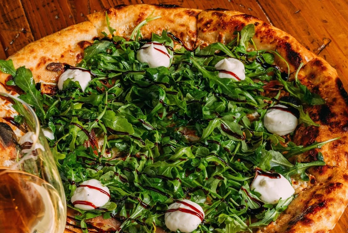 This photo shows a pizza topped with arugula, ricotta, prosciutto, dates and other ingredients.