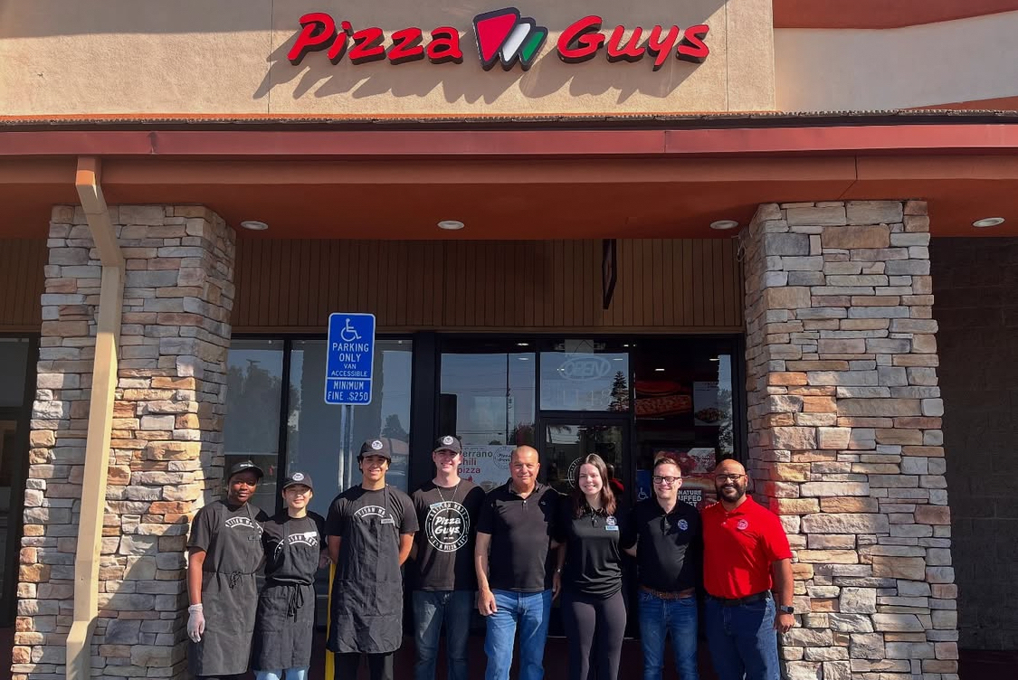 This photo shows a group of owners and employees standing in front of a new Pizza Guys location.