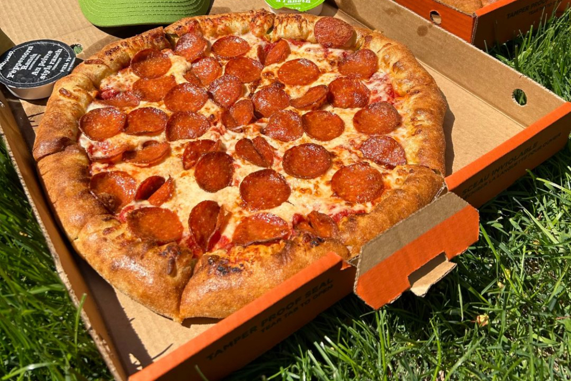 This photo shows a tasty-looking pepperoni pizza in a carryout box placed on green grass.