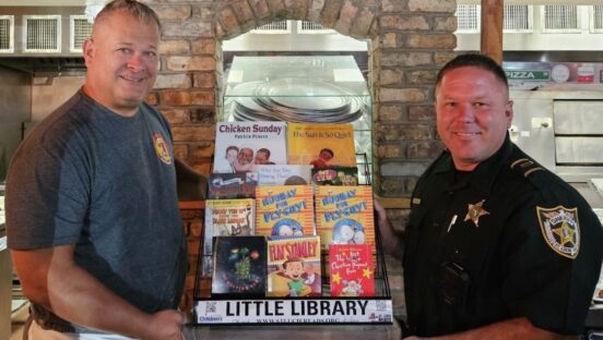 This photo shows Scott Van Duzer and a local police officer posing with a set of books in a display case in his pizzeria.
