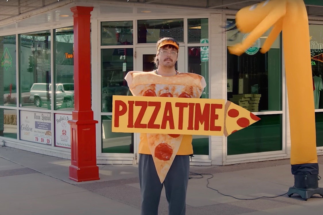 This screenshot from the video features band vocalist Austin Durry looking unhappy as he holds up a "PizzaTime" sign in front of a pizzeria.