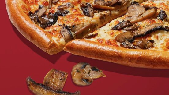 This photo shows a pizza topped with mushrooms and set against a dark red background, with two sliced mushrooms off to the side of the sliced pizza.