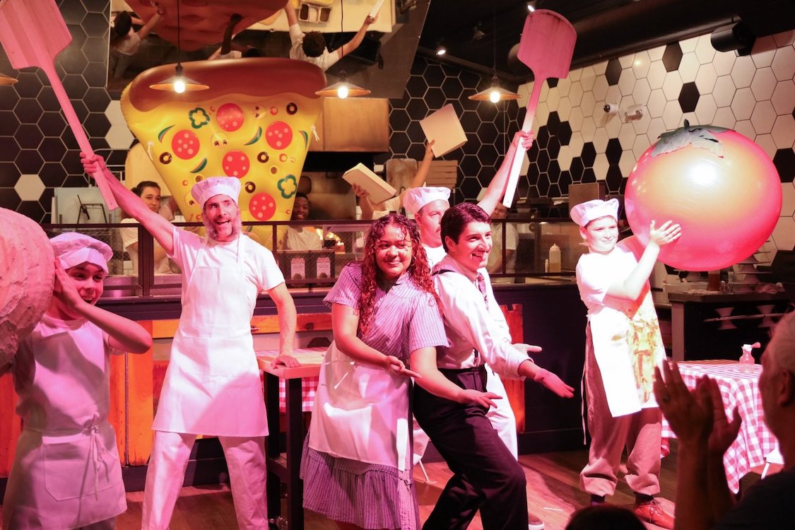 This photo shows a lively musical number from the play, with two actors extending their arms in dramatic fashion, two actors holding wooden peels in the air and a boy holding a large orange and red ball that appears to signify the planet Earth.