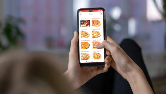 Woman holds a close-up of a mobile phone with a pizzeria website on the screen while lying on a sofa in a room.