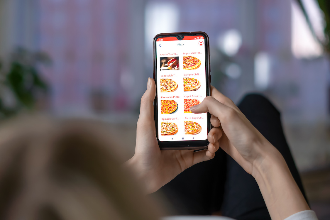 Woman holds a close-up of a mobile phone with a pizzeria website on the screen while lying on a sofa in a room.