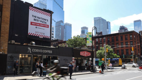 This photo shows a billboard in New York City touting that reads "The Nation's Best Pizza - Not You, New York." It's placed above a coffee shop. New Yorkers are seen walking by while, in the foreground, a delivery person speeds past on a bicycle in a blur.