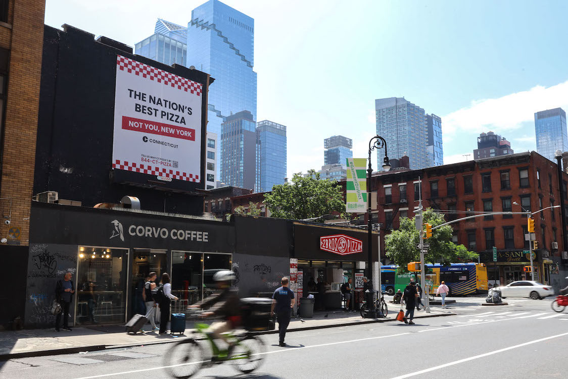 This photo shows a billboard in New York City touting that reads "The Nation's Best Pizza - Not You, New York." It's placed above a coffee shop. New Yorkers are seen walking by while, in the foreground, a delivery person speeds past on a bicycle in a blur.