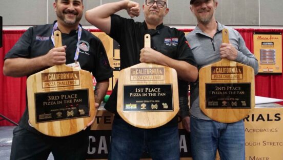 This photo shows three male members of the U.S. Pizza Team holding up their peel-shaped trophies at a previous California Pizza Challenge event. The man in the middle is humorously flexing the biceps of his right arm.