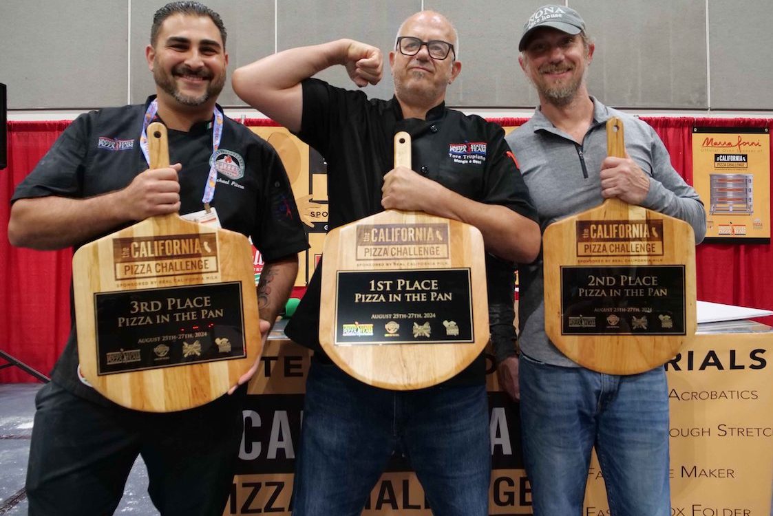 This photo shows three male members of the U.S. Pizza Team holding up their peel-shaped trophies at a previous California Pizza Challenge event. The man in the middle is humorously flexing the biceps of his right arm.